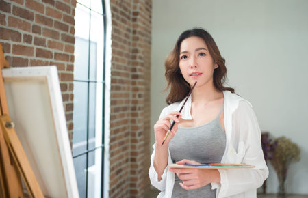 Portrait of a young woman artist is holding a brush and mix color oil painting on palette and thinking about something paint with canvas in house studioの写真素材