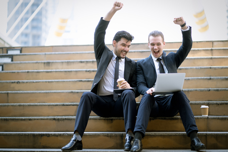 Two cheerful business man sitting on stairs celebrate success looking at laptop in urban city.Successful corporate team, winners excited of victoryの写真素材