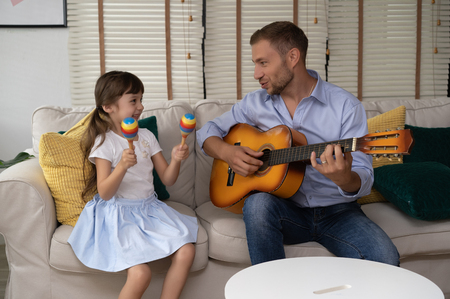 Happy Father's day.Funny portrait of smiling father playing guitar and her daughter holding maracas sitting sofa at homeの写真素材