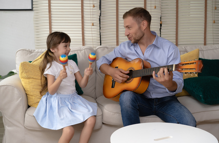 Happy Father's day.Funny portrait of smiling father playing guitar and her daughter holding maracas sitting sofa at homeの写真素材