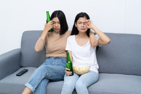 Funny two women sitting on couch watching sad movie depressed with sitting in living room sofa eating popcorn and beer.の写真素材