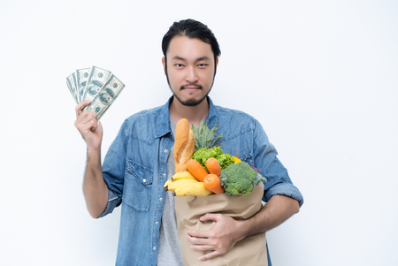 Happy young Asian male shopper, hands holding shopping bags full of groceries and show dollar bills, isolated standing on white background.の写真素材