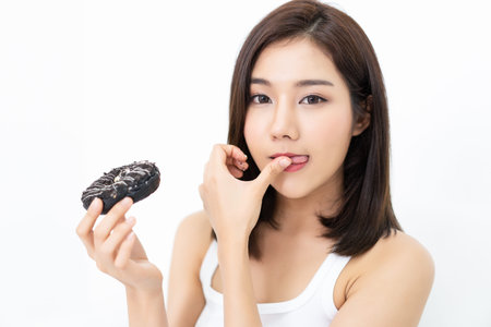 Beautiful Asian girl enjoys eating a donut with chocolate and sucking her finger on isolated on...
