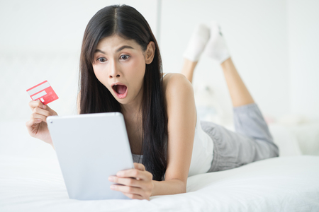 Amazed Asian woman using tablet and holding credit card her seeing big sales announcement on website while surfing internet lying on a bed of an her room. Shopping online, shocked female receiving unexpected email or messageの写真素材