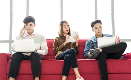 Students are sitting on the red sofa in university atrium social networking using several tech...