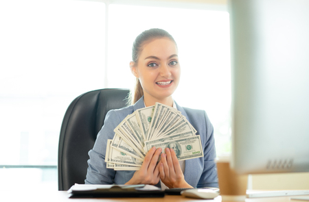 Happy brunette woman  with many dollars near laptop in office.の写真素材