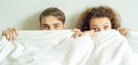 Funny couple lying in bed with blanket pulled up over their noses and staring at camera with surprised and shocked look.Caucasian models in love, relationship, dating, happy people ,bedtime, love, romance and sex concept.Horizontal banner compositionの写真素材