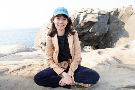 Portrait of happy young Asian woman sitting on top of a rock a cliff view tropical beautiful sea and looking at camera at the Elephant trunk rock at the north coast of Taiwan, Shenao, New Taipei City, Taiwan.の写真素材
