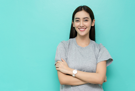 Portrait of happy beautiful young Asian woman in gray t-shirt standing with crossed arms and smiling on green pastel backgroundの写真素材