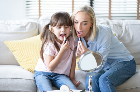 Beautiful mom with her little cute daughter are doing your makeup and having fun. Mom and daughter wear lipstick look at mirror sitting at sofa in living room.People and family and lifestyle mother's day conceptの写真素材
