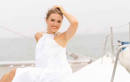Portrait of a beautiful woman in white dress sitting on the side of a sail boat enjoying mild sunlight and looking at camera. Lifestyle, freedom and vacation concept.の写真素材