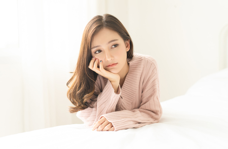 Woman indoor portrait. Closeup of pensive young Asain woman in warm knitted pink clothes in the bedroom. Negative human facial expression.の写真素材
