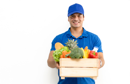 Handsome Caucasian grocery delivery courier man in blue uniform with grocery wooden box with fresh fruit and vegetable. Shopping service concept, isolated standing on white background.の写真素材