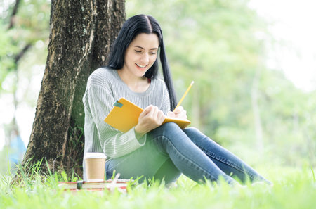 Smiling student girl sitting in a park. College girl making notes in a book, preparing for exams...