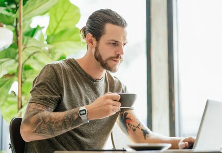 Concentrated hipster man with bearded working on laptop and drinking coffee or tea at coffee shop. Online marketing, education, e-learning.の写真素材