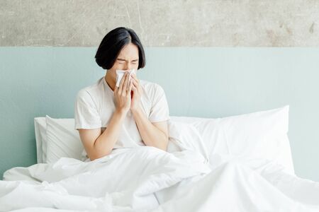 Sick young Asian man blowing his nose while sitting on bed at home.の写真素材