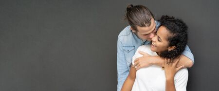 Panoramic banner image of a sweet couple hugging and kissing his wife on the cheek on black background in studio. wide cropの写真素材