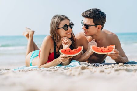 Happy young couple holding and eating slices of watermelon on the beach.Casual lifestyle, love, dating, Vacation concept.の写真素材