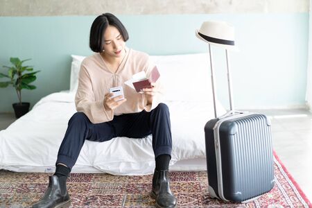 Happy young traveler Asian man sitting on bed in hotel room accomodation while checking his flight online for holiday. Technology, People travel and lifestyle conceptの写真素材