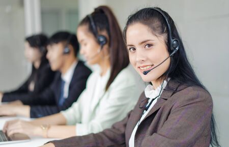 Beautiful young woman with headsets consulting client on phone in customer support service while colleagues in background at office and looking at camera. Operator service business representative concept.の写真素材