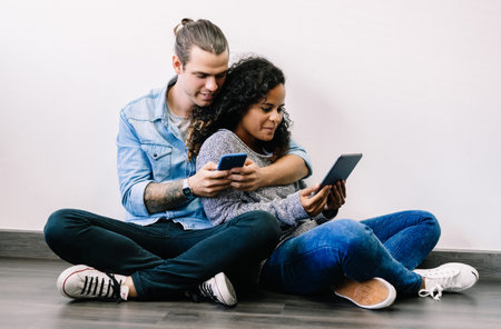 Attractive happy young multiethnic couple sitting on the floor using smartphone and tablet at home....