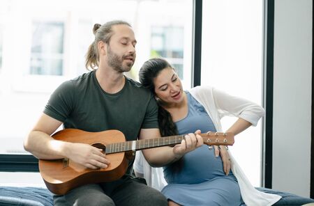 Loving in a living room. Handsome husband and his beautiful pregnant wife plays the guitar, sings the songs together on the sofa at home. Pregnant Woman at Home Concept. Happy family people.の写真素材