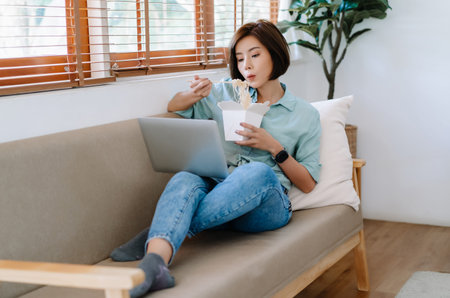 Portrait of young Asian woman freelancer eating chinese food from delivery while working on laptop...