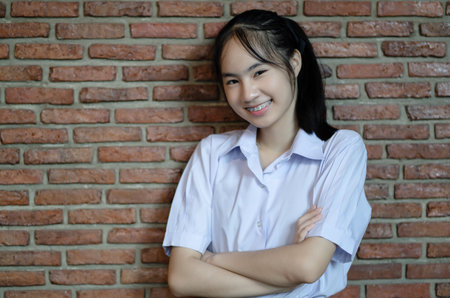 Portrait of smiling teenager in uniform standing with arms crossed at red brick wall and looking at...