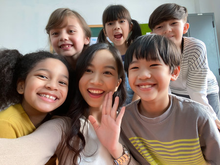 Smiling young Asian teacher making selfie with her schoolchildren in classroom. Elementary...