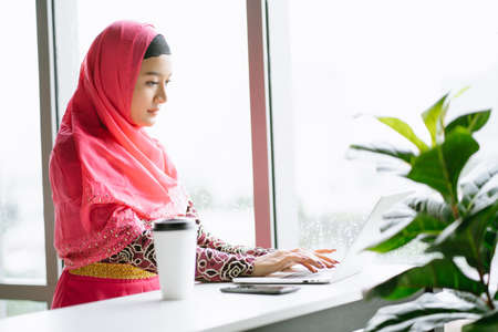 Portrait of young Muslim woman in hijab pink dress using laptop computer sitting at cafe table. Muslim business professional conceptの写真素材