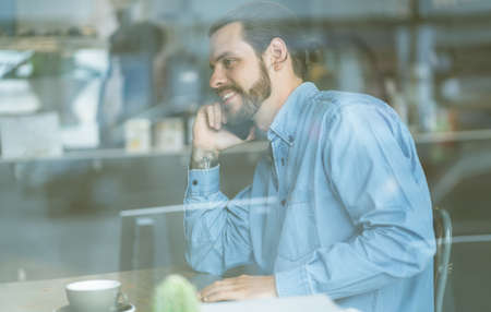 Stylish male freelancer talking on the mobile phone and smiling while sitting at table behind glass in cafe.Cozy office workplace.の写真素材