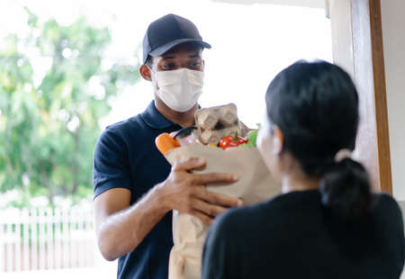 African American delivery man with medical mask delivering food to a woman at home.Online shopping and Express delivery. Under quarantine, disease outbreak, coronavirus covid-19 pandemic.の写真素材