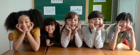 Portrait of happy elementary international students sitting on desk touches cheeks with both hands and looking at camera in classroom.Web banner panorama with adorable school kids.Education concept.の写真素材