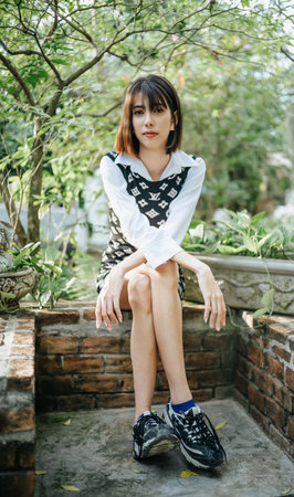 Portrait of young Asian woman sitting on stone border and looking at camera in sunny park. Holiday activity and lifestyle concept.のeditorial素材