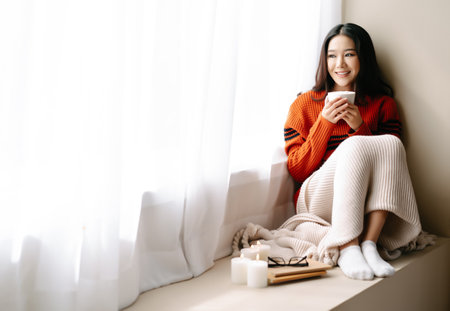 Young beautiful happy Asian woman wearing knitted warm sweater sitting home in the chair by the window with cup of hot drink at home.Cozy winter decorated with candles.Feeling comfortable at home.の写真素材