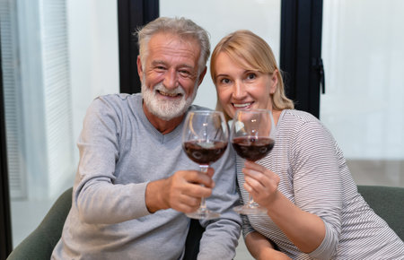 Senior romantic couple sitting together at home smiling and drinking wine and enjoy in love, looking at camera. Retired elderly joy lifestyle concept.の写真素材