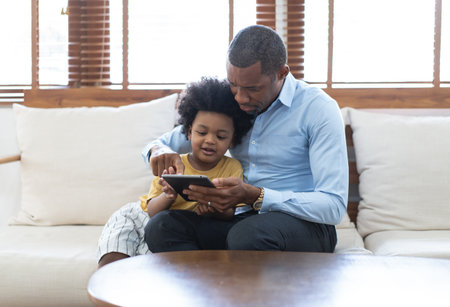 Portrait of happy African American father and son using a tablet while sofa sitting on sofa at home. Having great time together. Concept of family entertainment, education, technology.の写真素材