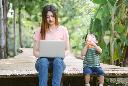 Portrait of young Asian mother and little son sit together on the floor in garden while mom working remotely on laptop and son playing with soap bubbles machine. Childcare and work concept.の写真素材