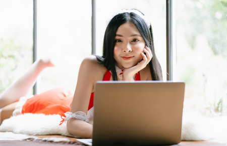 Beautiful young sexy Asian woman lying on carpet with laptop and smiling looking at camera at home. Young millennial generation  lifestyle and technology concept.の写真素材