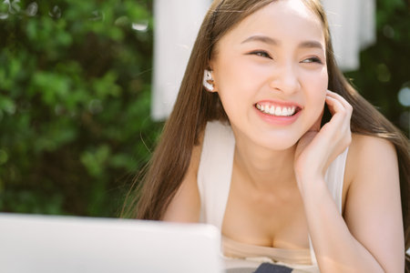 Closeup portrait of beautiful young Asian smilling lying on sofa patio furniture on outdoor patio living room while using laptop and wireless earphone for video call. Holiday and lifestyle concept.の写真素材