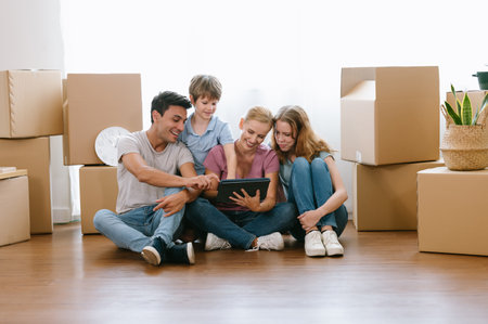 Happy caucasian family with teenage daughter and younger brother sitting on floor among cardboard boxes have fun using modern tablet buying furniture into new house.E-commerce retail services concept.の写真素材