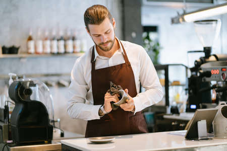 Portrait of handsome barista standing in behind the counter bar and smiling while wiping ceramic cups in a coffee shop. Clean equipment. 
Startup small business owner and Service mind concept.の写真素材