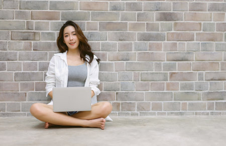 Portrait of happy Korean girl in casual sitting on floor with crossed legs while using laptop computer and looking at camera at copy space on brick wall background. Technology and lifestyle concept.の写真素材