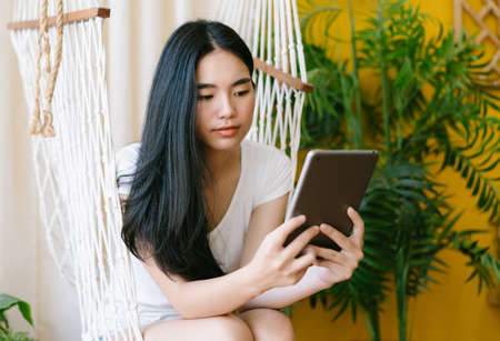 Portrait of young Asian woman reading a message, e-book or information on her tablet computer while sitting on swing in the living room. Social media time, modern tech easy and comfy usage concept.の写真素材