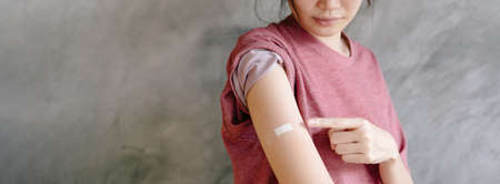 Young Asian woman showing arm after vaccine injection on loft concrete wall background, closeup, panorama with free space. Vaccination and health care during pandemic covid-19.の写真素材