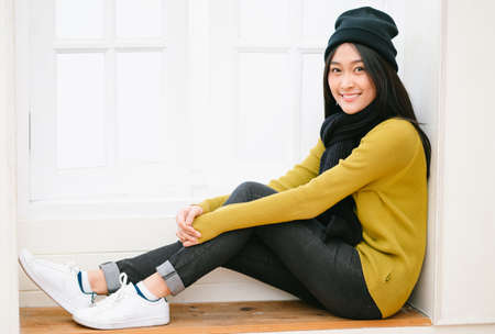 Young beautiful Asian woman sitting by the window and looking at camera. Lifestyle, comfort and Relax concept. Fashion, Autumn, winter.の写真素材
