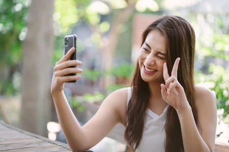 Portrait of cool cheerful Asian girl smile and showing peace sign while taking selfie photo on cellphone in the park. People sincere emotions lifestyle concept.の写真素材
