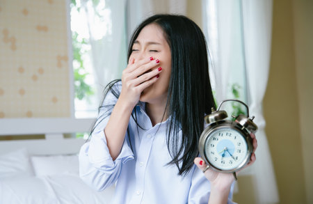Portrait of young sleepy Asian woman holding alarm clock after awakening and yawning in bed. Early...