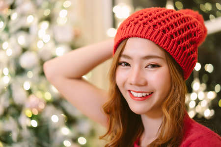 Closeup of beautiful natural looking young smiling Asian woman wearing knitted sweater and winter hat taking selfie in room house with newyear decoration indoors, background Christmas tree.の写真素材