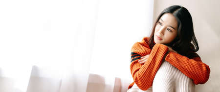 Beautiful young Asian woman wearing knitted warm sweater feeling depress sad girl lonely at home window thinking negative thoughts. Bad feelings stress, grief, emotions. Mental health banner panorama.の写真素材